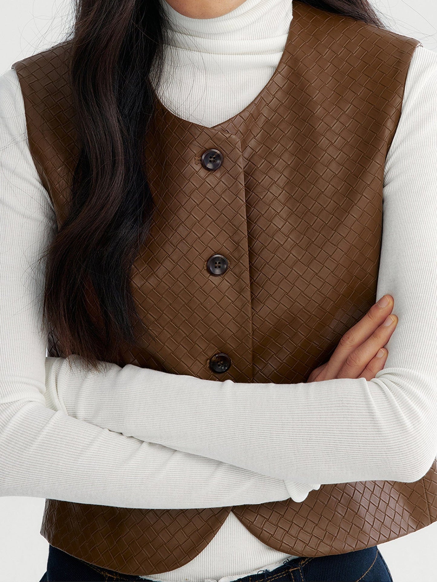 Close up of a woman wearing Arkin Studio artisan woven leather vest in brown with a white long sleeve top inside, and navy pants, focus on her both arms crossing over the chest, standing facing the camera on a white background