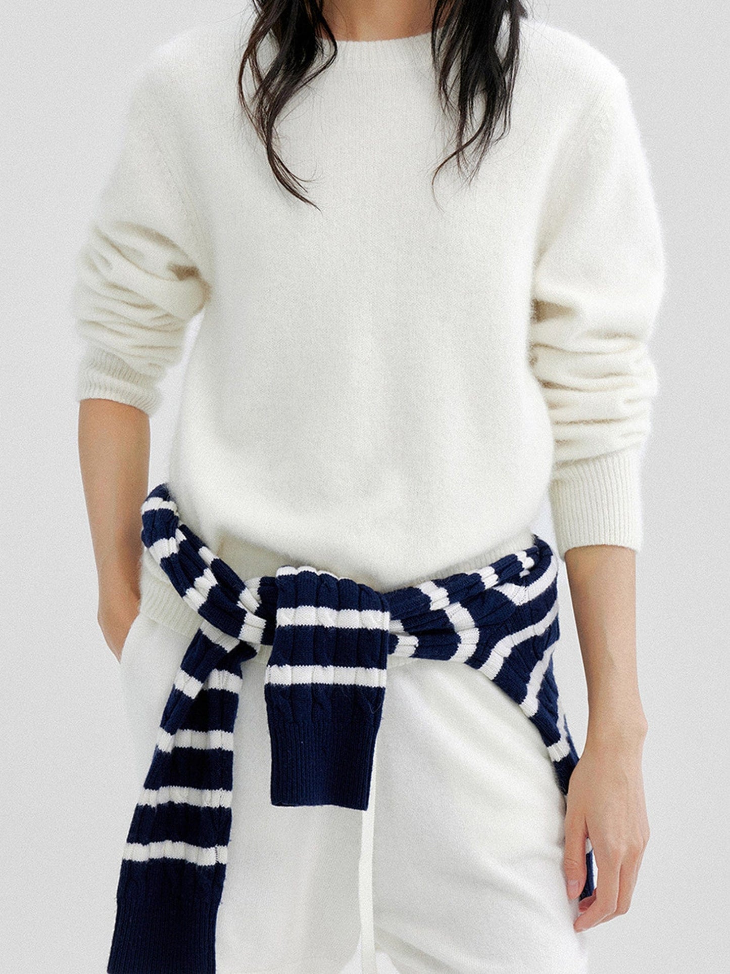 Woman wearing Arkin Studio cozy crewneck wool sweater in ivory and matching shorts, with a striped knitwear around waist, standing facing the camera, on a white background