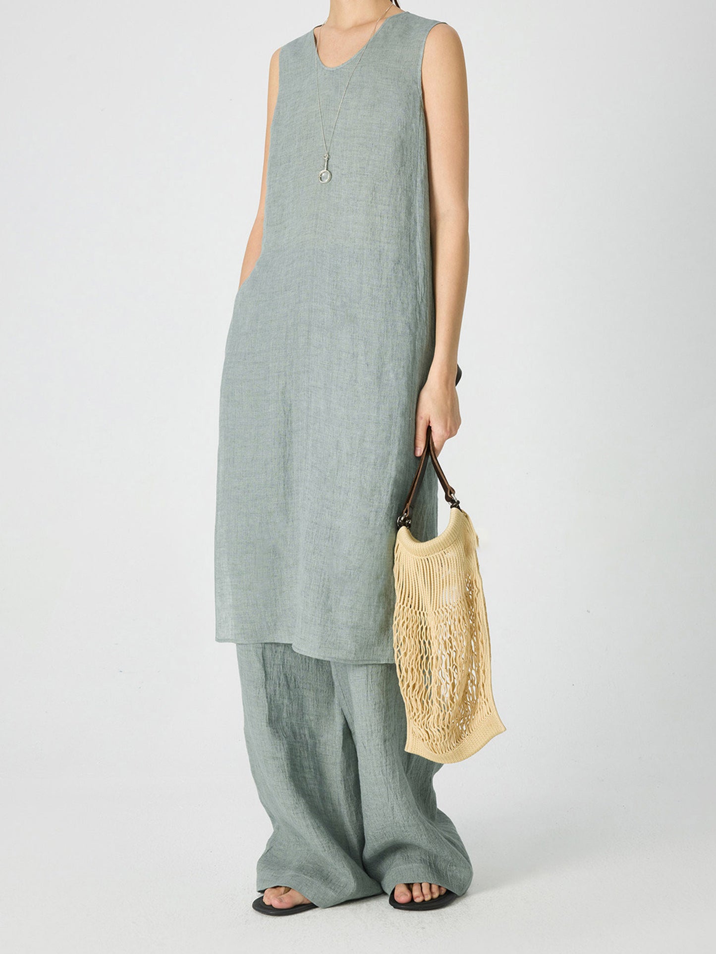 Woman wearing Arkin Studio crinkled linen wide leg pants in mint-green, with a pair of sandals, holding a yellow woven bag, standing sideways, on a white background
