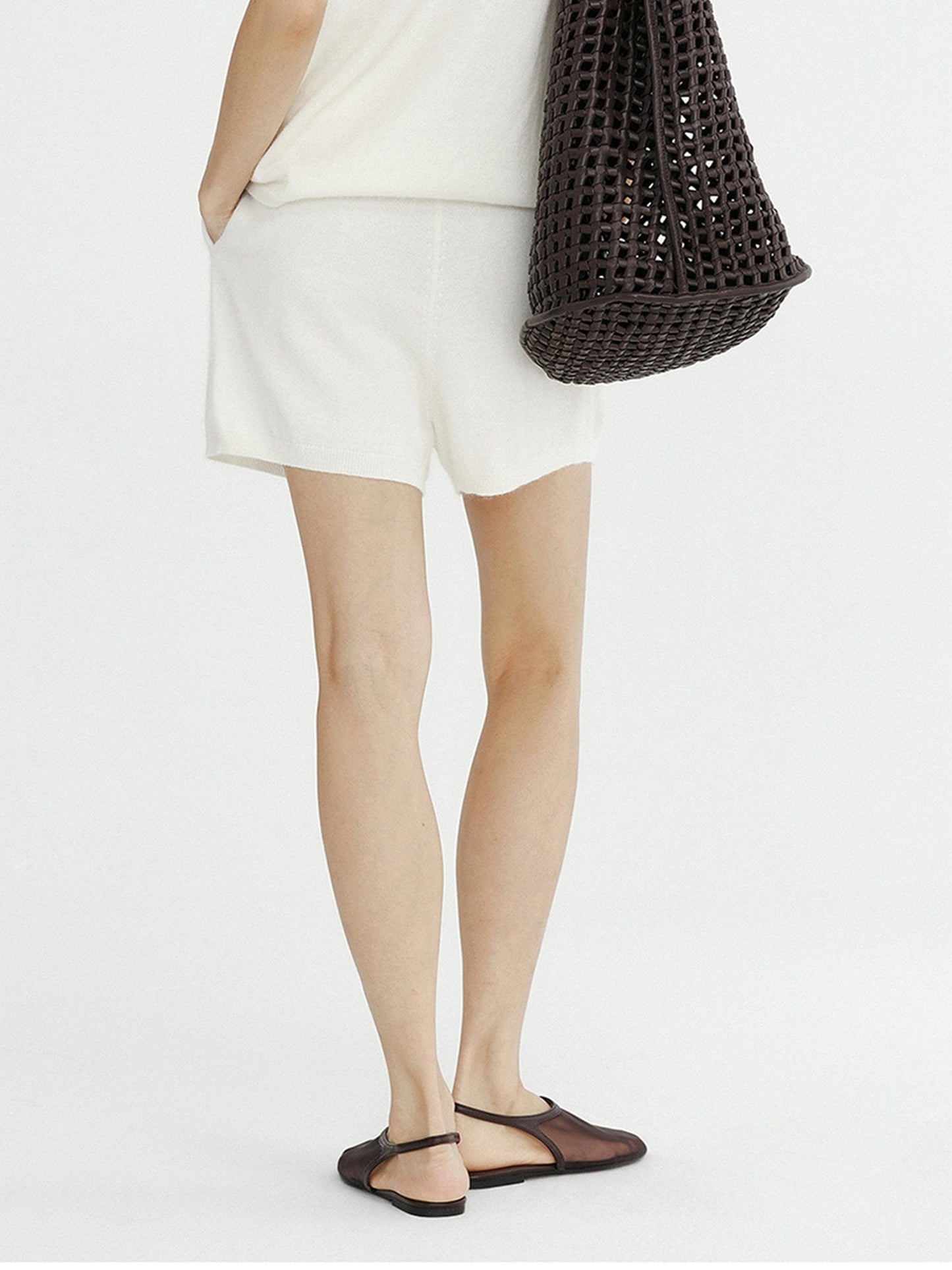 Woman wearing Arkin Studio ribbed knit drawstring shorts in white and matching top, wearing a pair of chocolate shoes, a chocolate bag on her right shoulder, standing facing a white wall, on a white background