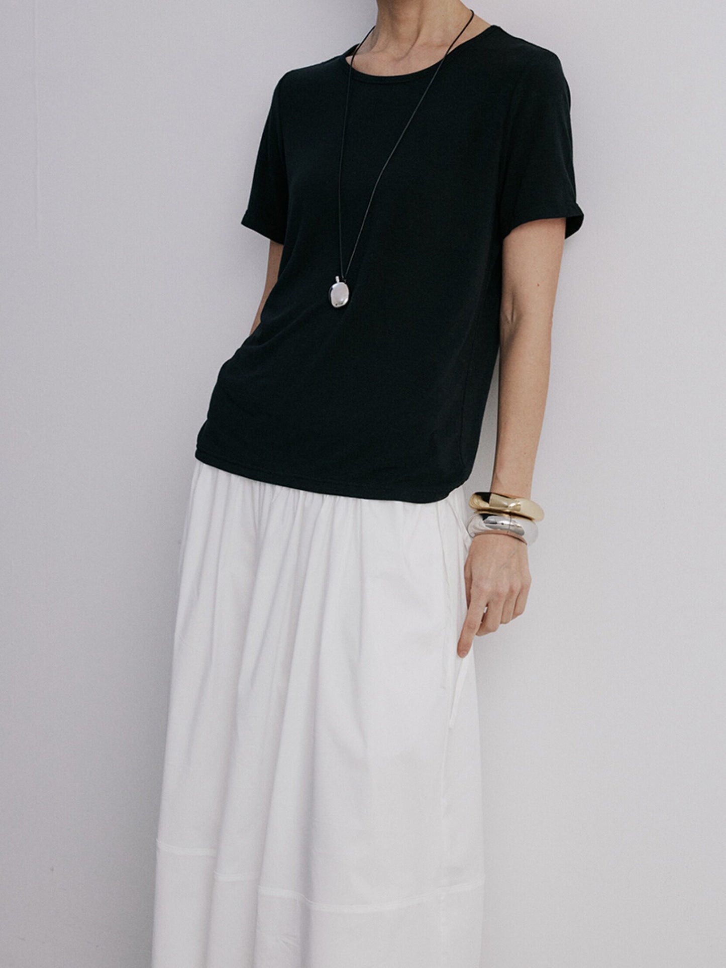 Woman wearing Arkin Studio lightweight pleated a-line drawstring skirt in white and a black t-shirt, leaning against a wall, on a white background