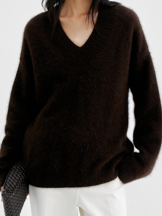 Woman wearing Arkin Studio oversized v-neck fuzzy sweater in dark-brown and matching white pants, holding a black bag, standing facing the camera, on a white background