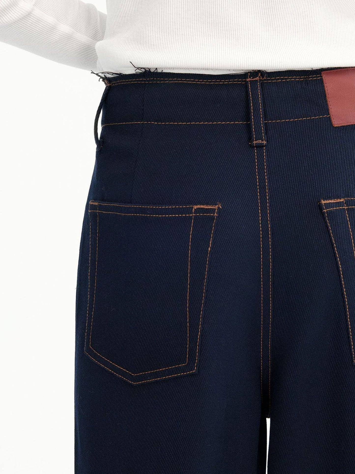 Close-up of the back and hip area of a woman wearing Arkin Studio dark navy raw edge contrast stitch trousers and a white long-sleeve tee, standing with back to camera, focusing on the pants' back design, on a white background
