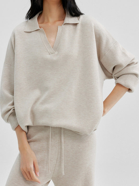 Woman wearing Arkin Studio relaxed v-neck wool blend sweater in taupe and matching pants, one hand behind her back and another on her leg, standing facing the camera, on a white background