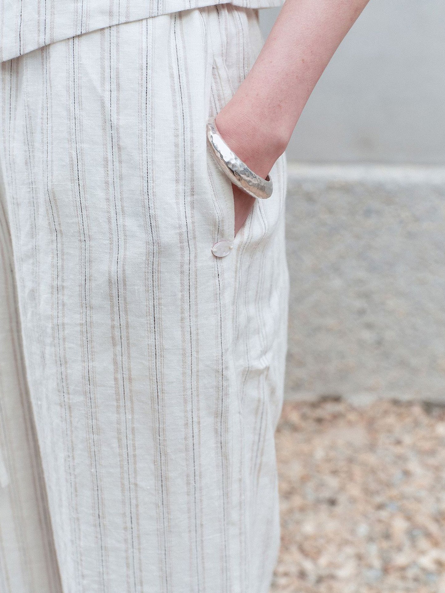 Close-up of a woman's hand tucked into the pocket of Arkin Studio striped linen drawstring wide leg pants in ivory, matching vest, standing on a stone floor