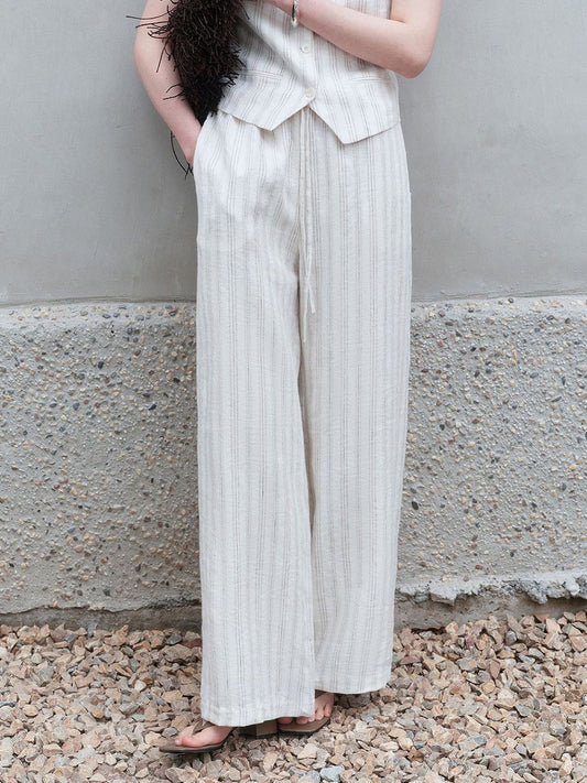 Woman wearing Arkin Studio striped linen drawstring wide leg pants in ivory and matching vest, with a brown flat bag tucked under her arm, standing on a stone floor, facing the camera