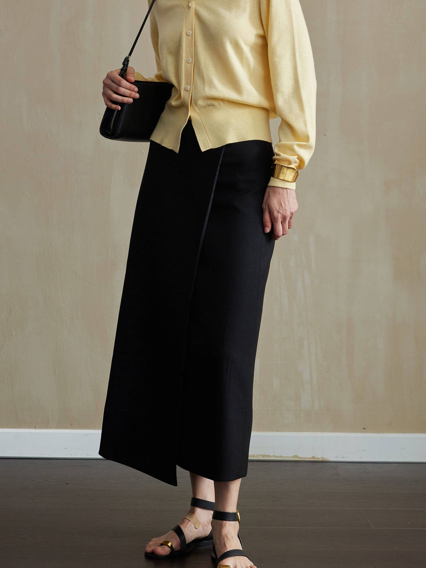 Woman wearing Arkin Studio tencel wool twill a-line skirt in black and a yellow cardigan, with summer sandals on feet, slightly turned to the side, on the dark-brown floor, a yellow wall behind her