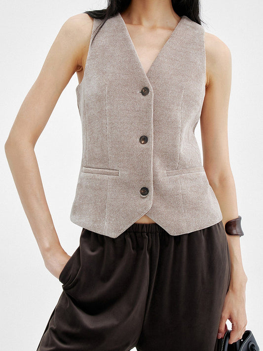 Woman wearing Arkin Studio v-neck corduroy vest in taupe and brown pants, left hand holding a black bag and right hand in a pocket, standing facing the camera, on a white background