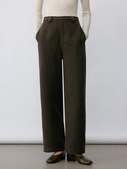 Woman wearing Arkin Studio wool blend straight leg trousers in olive-green and a white long sleeve top, wearing a pair of brown shoes, with hands in the pockets, standing facing the camera in front of a white wall, on a khaki floor