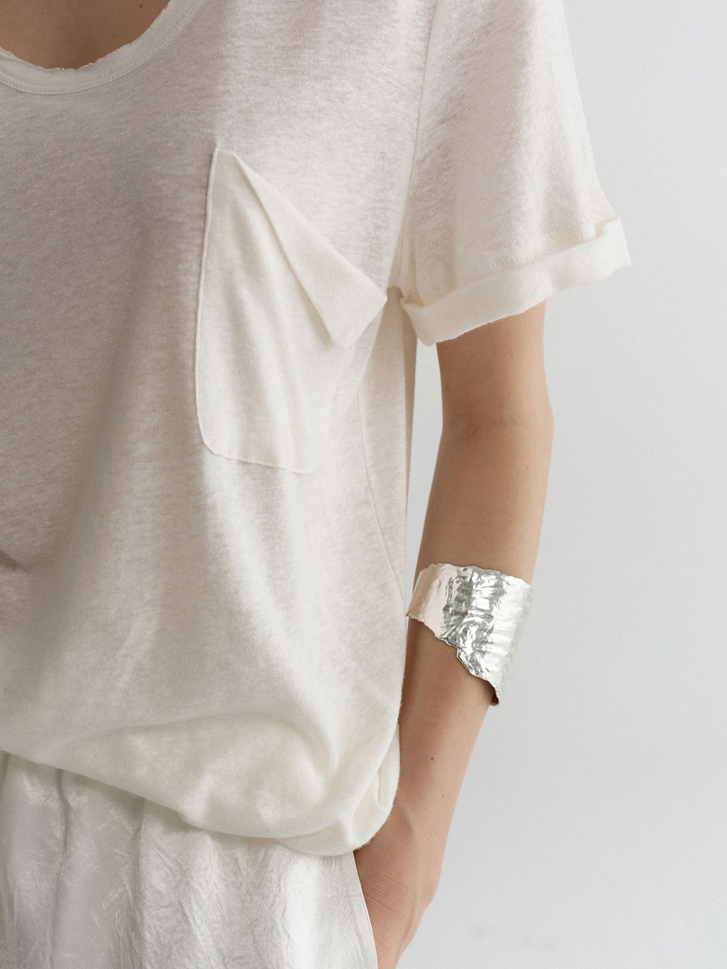Close-up of a woman's left upper body and chest area, wearing Arkin Studio airy u-neck linen tee in ivory and matching pants, one hand in the pants' pocket, facing the camera, on a white background