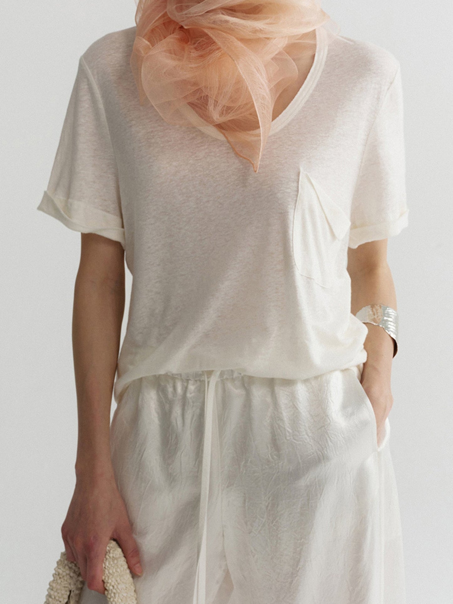 Woman wearing Arkin Studio airy u-neck linen tee in ivory and matching pants, with a delicate light pink sheer scarf wrapped around her neck, holding a white woven handbag, one hand in the pants' pocket, standing facing the camera, on a white background