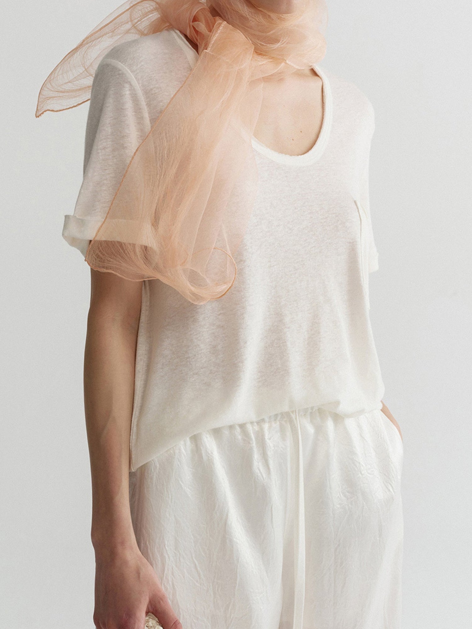 Woman wearing Arkin Studio airy u-neck linen tee in ivory and matching pants, with a delicate light pink sheer scarf wrapped around her neck, one hand in the pants' pocket, standing sideways,  on a white background