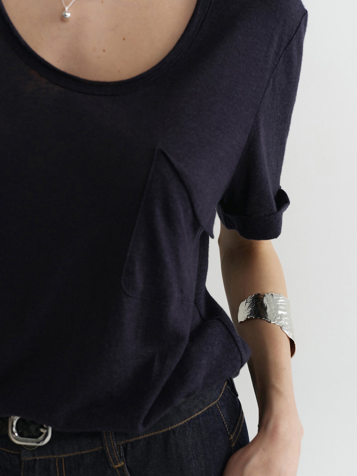 Close-up of a woman's left upper body and chest area, wearing Arkin Studio airy u-neck linen tee in navy and matching pants, one hand in the pants' pocket, facing the camera, on a white background