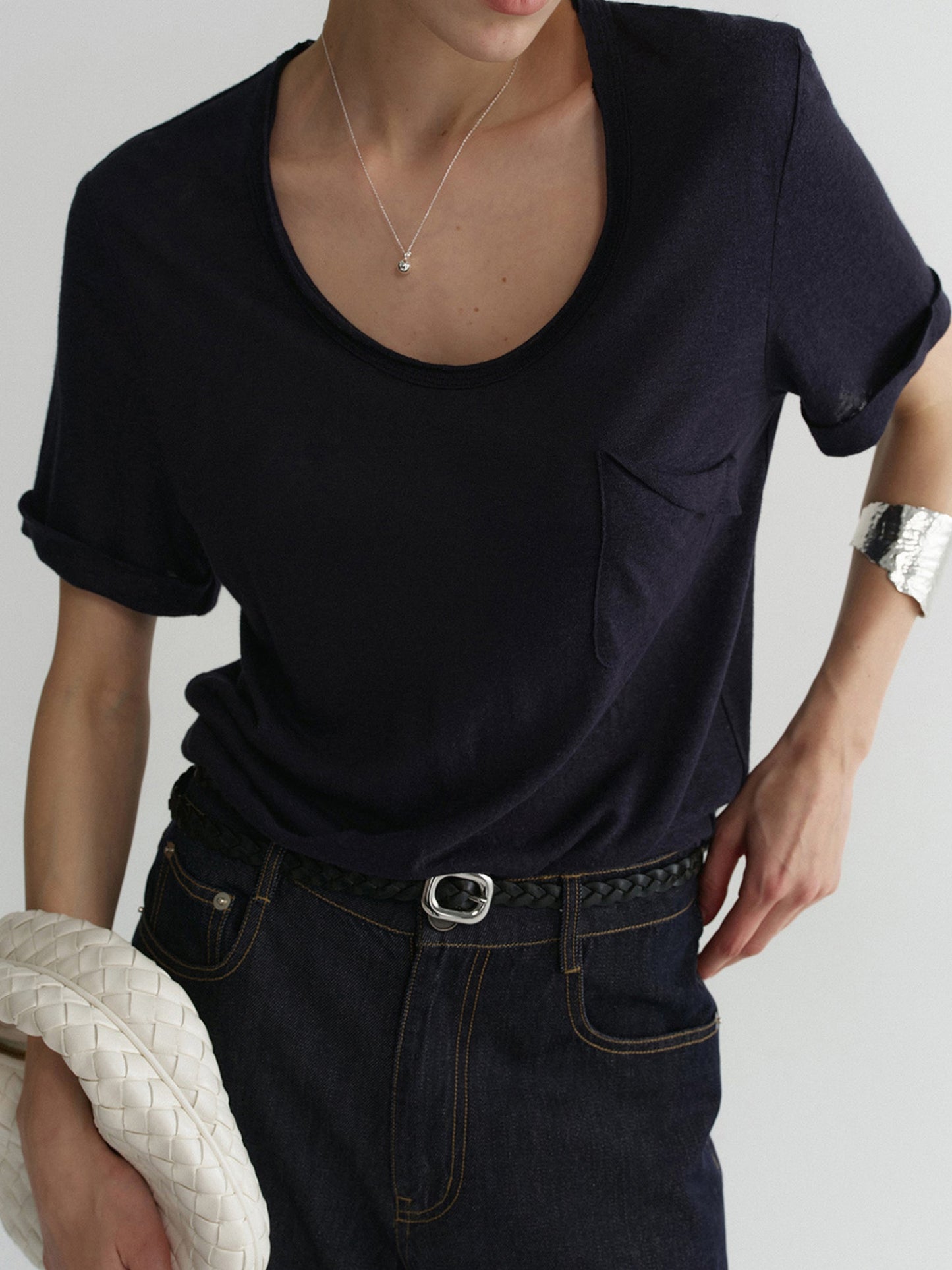 Woman wearing Arkin Studio airy u-neck linen tee in navy and matching pants, holding a white woven handbag in right hand, the left hand is touching the pants, standing on a white background