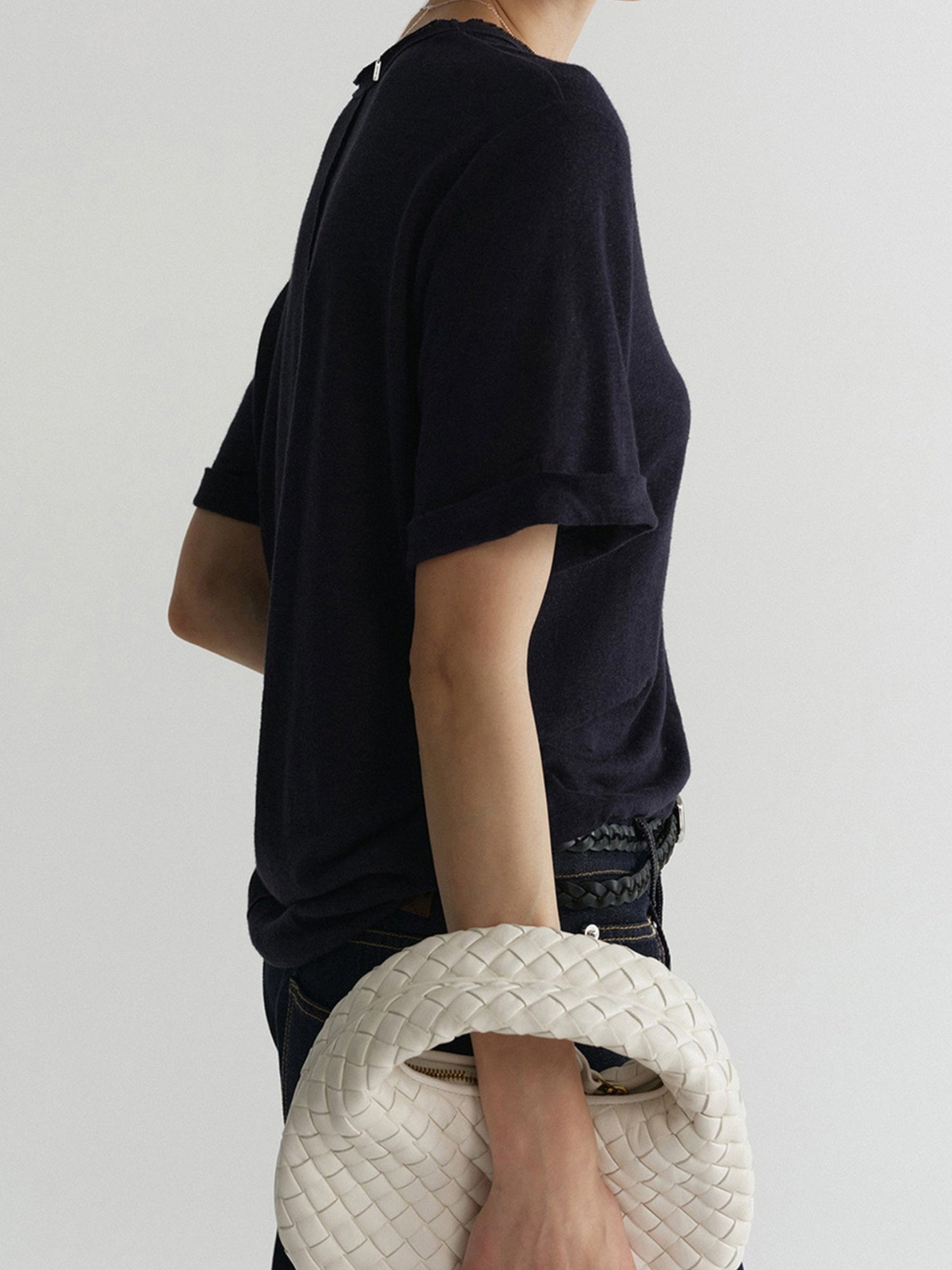 Woman wearing Arkin Studio airy u-neck linen tee in navy and matching pants, holding a white woven handbag in right hand, standing sideways, on a white background