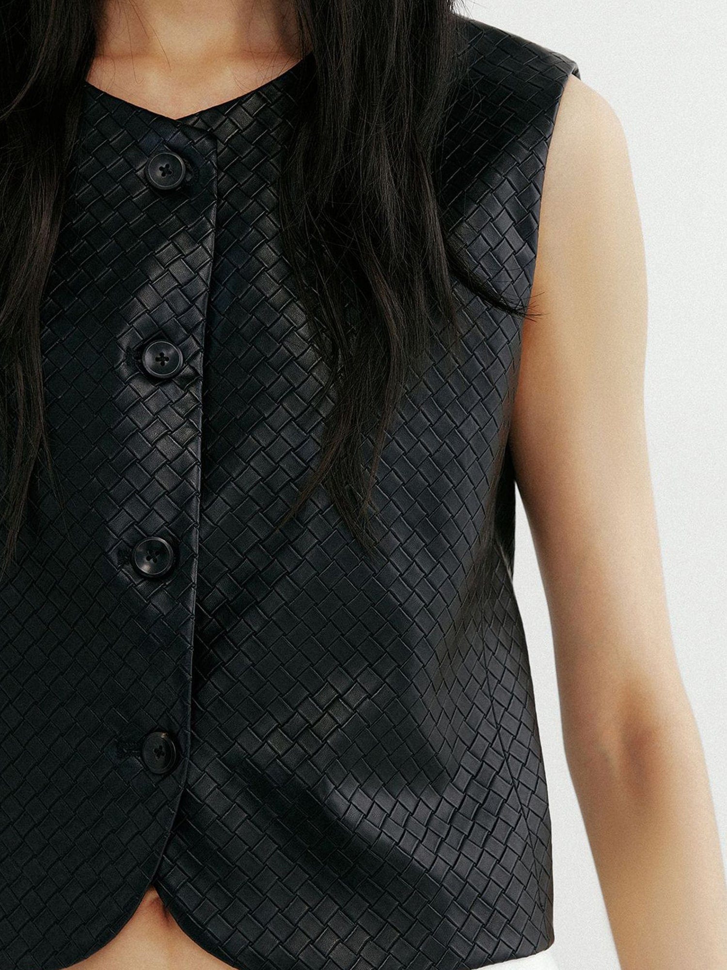 Close up of a woman wearing Arkin Studio artisan woven leather vest in black, one hand down at her one side, focus on her chest, standing facing the camera on a white background