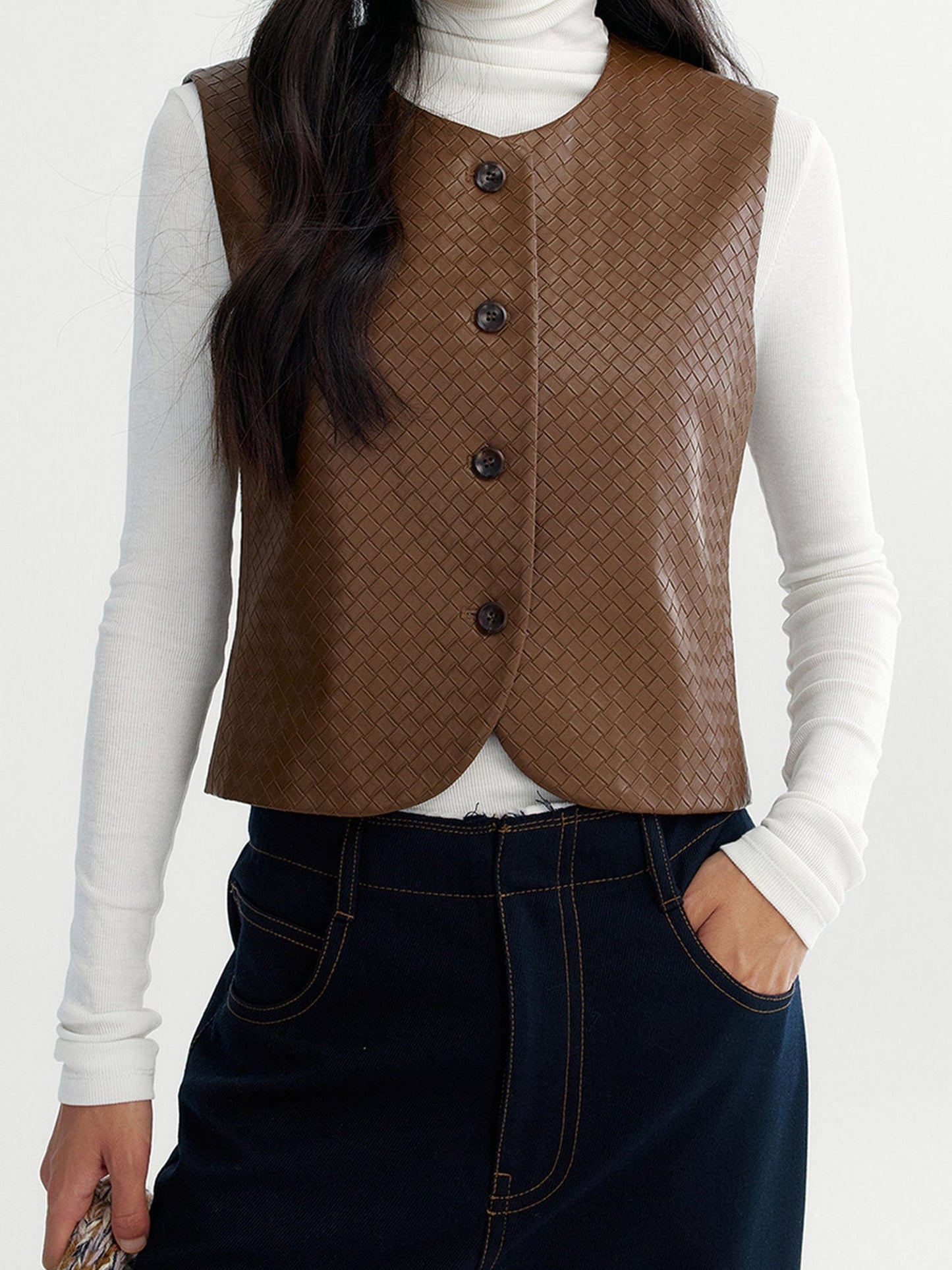 Woman wearing Arkin Studio artisan woven leather vest in brown with a white long sleeve top inside, and navy pants, one hand down at her side, another in her pocket, standing facing the camera on a white background