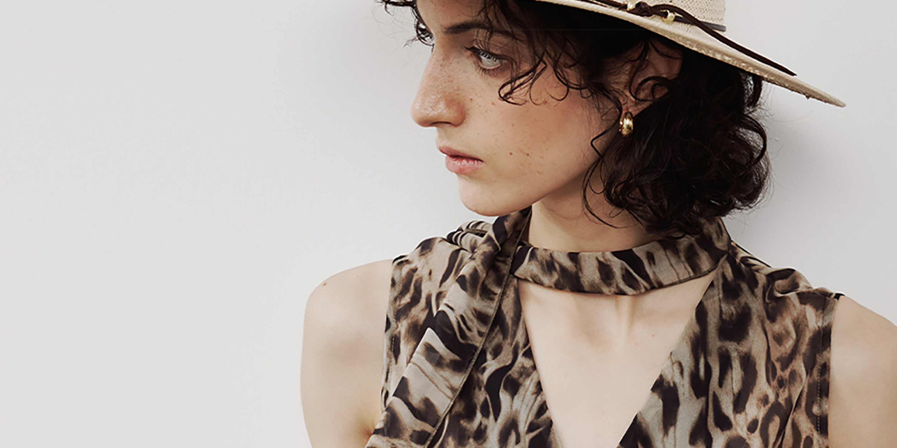 Woman wearing Arkin Studio leopard print sleeveless top and a wide-brim straw hat, short curly hair, gazing to the side, home banner - shop the collection