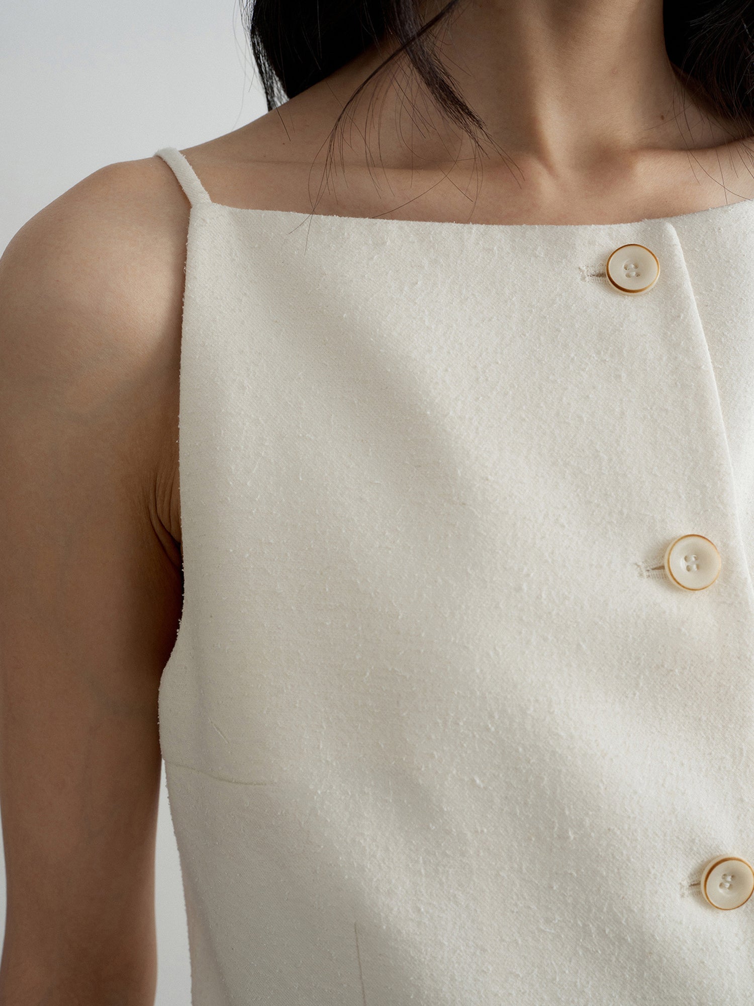 Close-up of the right chest area of a woman wearing Arkin Studio boatneck slim strap button front vest in ivory, standing facing the camera, on a white background