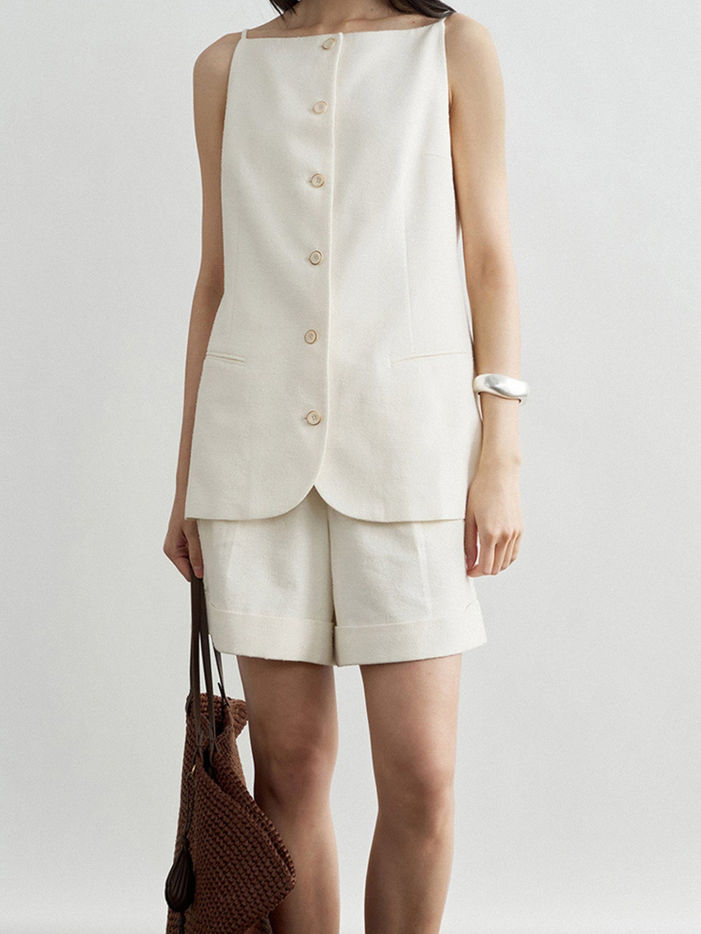 Woman wearing Arkin Studio boatneck slim strap button front vest in ivory and matching shorts, holding a woven bag, with a silver bracelet on her left hand, standing facing the camera, on a white background