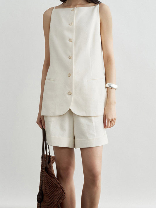 Woman wearing Arkin Studio boatneck slim strap button front vest in ivory and matching shorts, holding a woven bag, with a silver bracelet on her left hand, standing facing the camera, on a white background