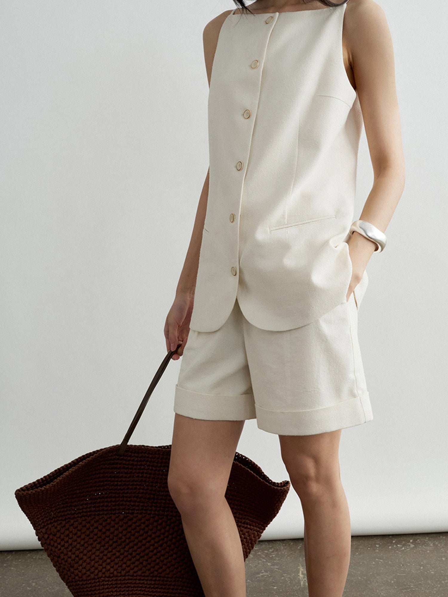 Woman wearing Arkin Studio boatneck slim strap button front vest in ivory and matching shorts, holding a woven bag, with a silver bracelet on her left hand, standing sideways on the floor, in front of a white background
