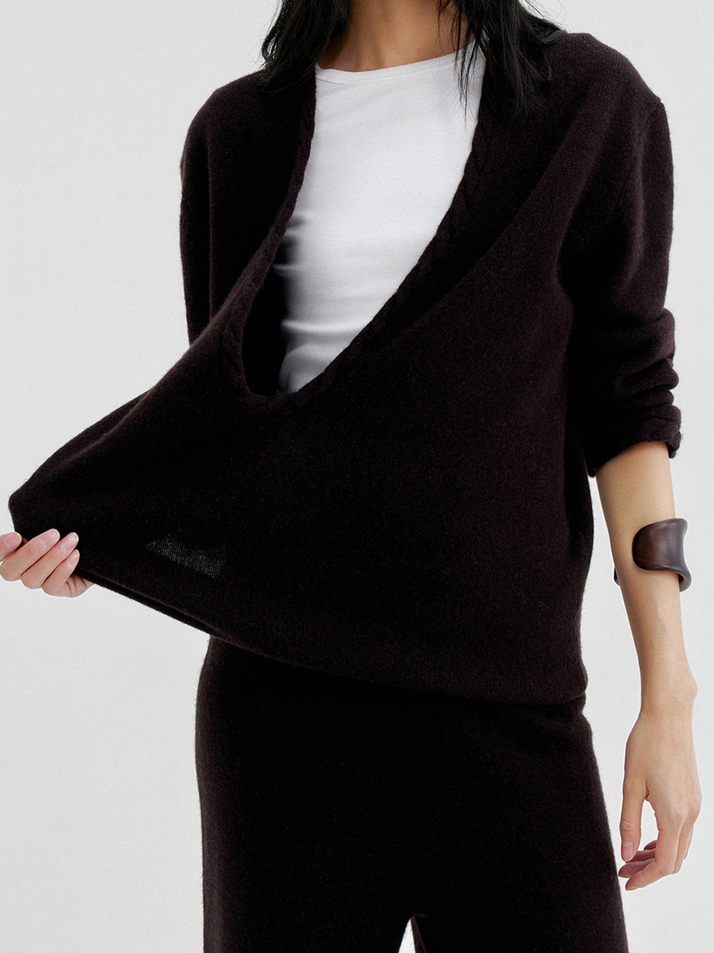 Woman wearing Arkin Studio cable knit v-neck wool sweater in dark-brown with a white top inside, matching knit pants, one hand pulling the sweater, standing facing the camera, on a white background