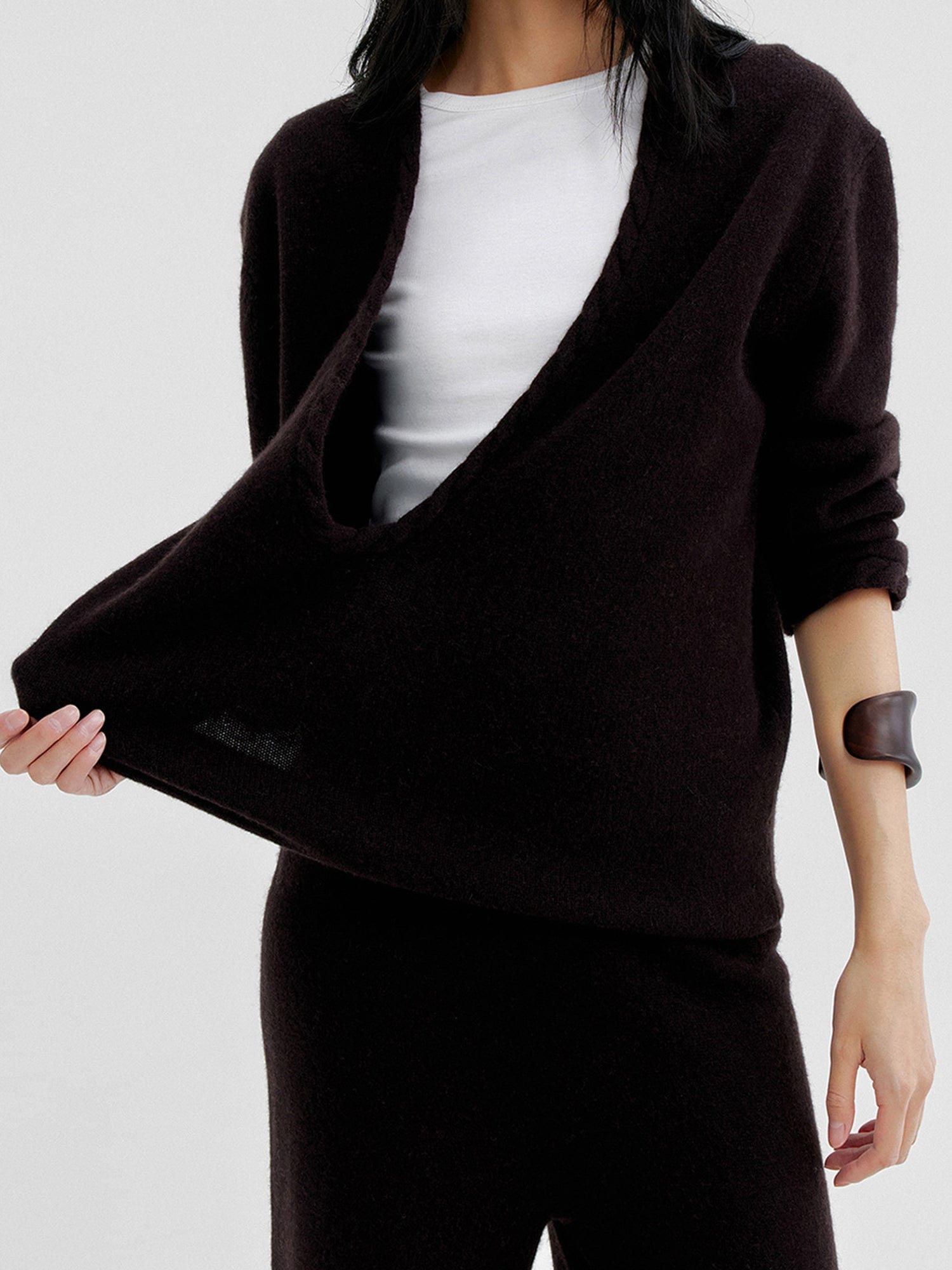 Woman wearing Arkin Studio cable knit v-neck wool sweater in dark-brown with a white top inside, matching knit pants, one hand pulling the sweater, standing facing the camera, on a white background