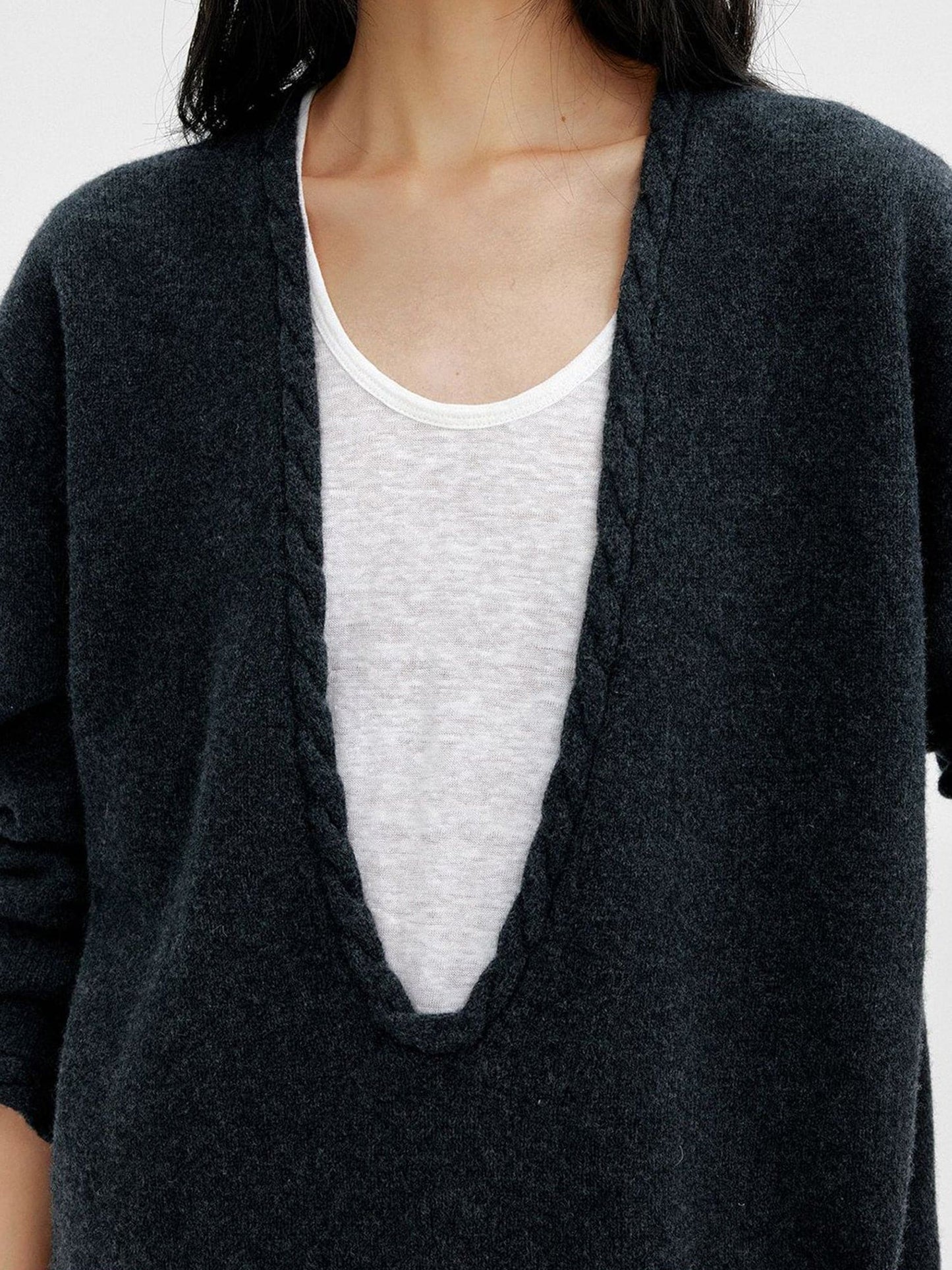 Close up of a woman wearing Arkin Studio cable knit v-neck wool sweater in dark-gray with a white top inside, focus on the upper part of her, standing facing the camera, on a white background