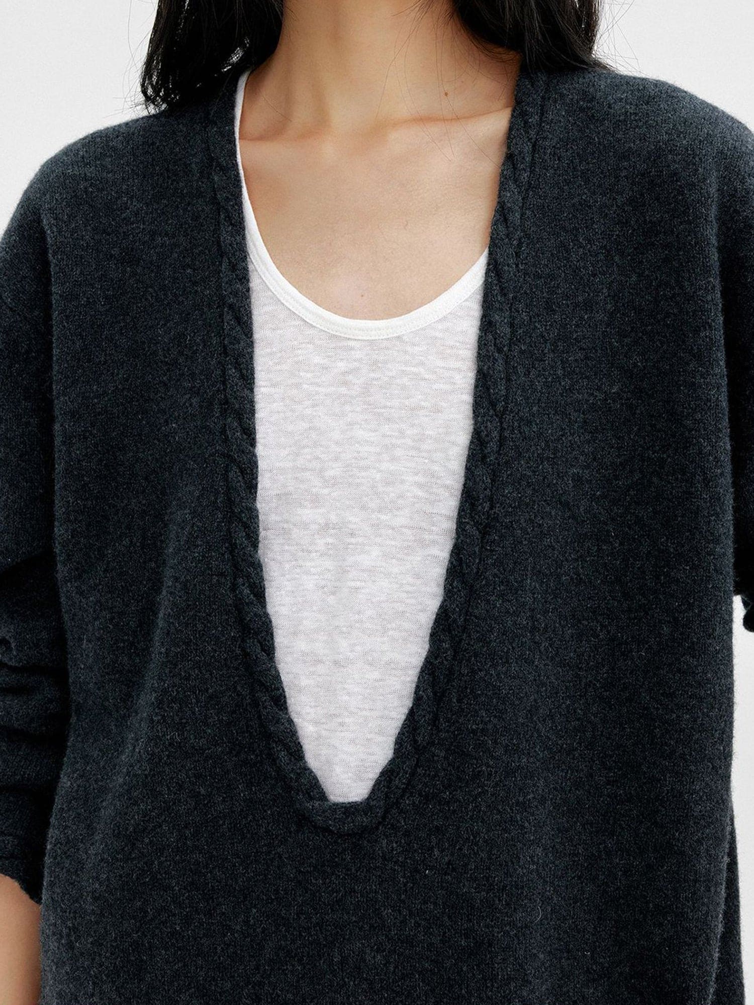 Close up of a woman wearing Arkin Studio cable knit v-neck wool sweater in dark-gray with a white top inside, focus on the upper part of her, standing facing the camera, on a white background