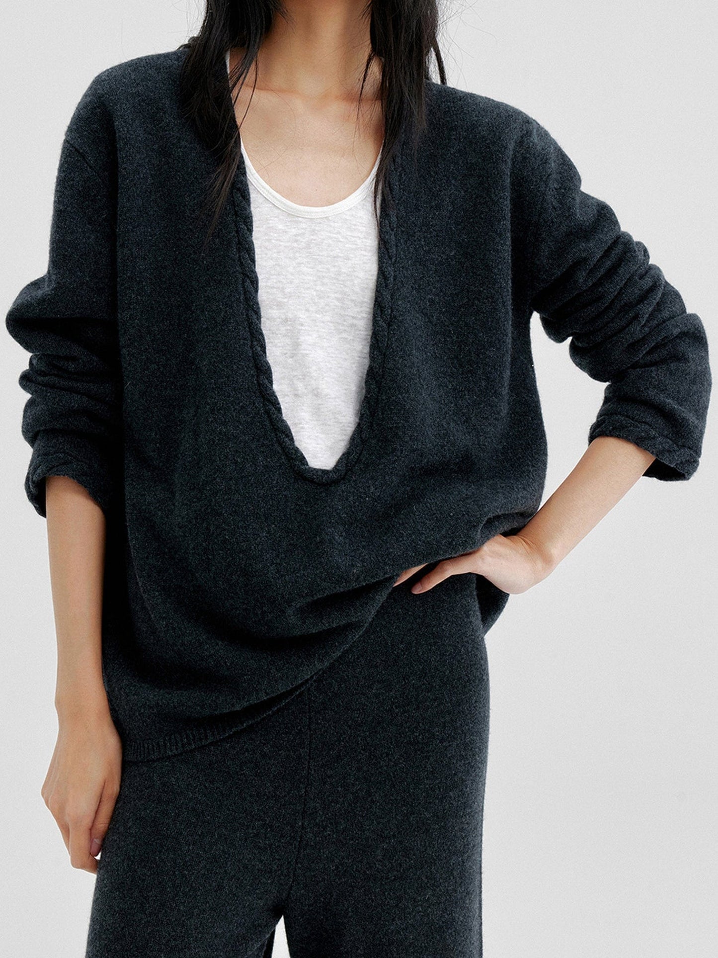 Woman wearing Arkin Studio cable knit v-neck wool sweater in dark-gray with a white top inside, matching knit pants, one hand touching the waist, standing facing the camera, on a white background