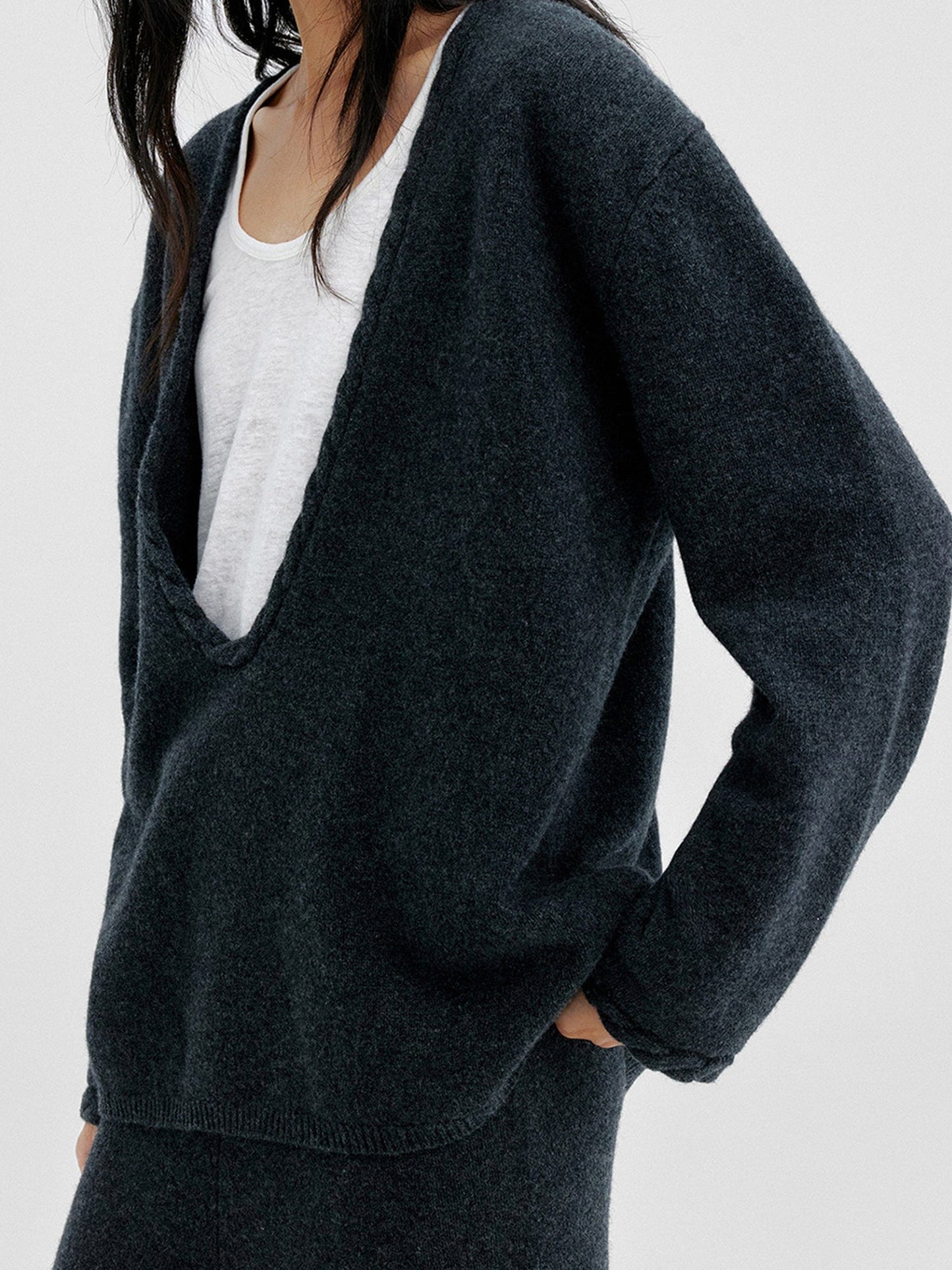 Woman wearing Arkin Studio cable knit v-neck wool sweater in dark-gray with a white top inside, matching knit pants, one hand in a pocket, standing sideways, on a white background