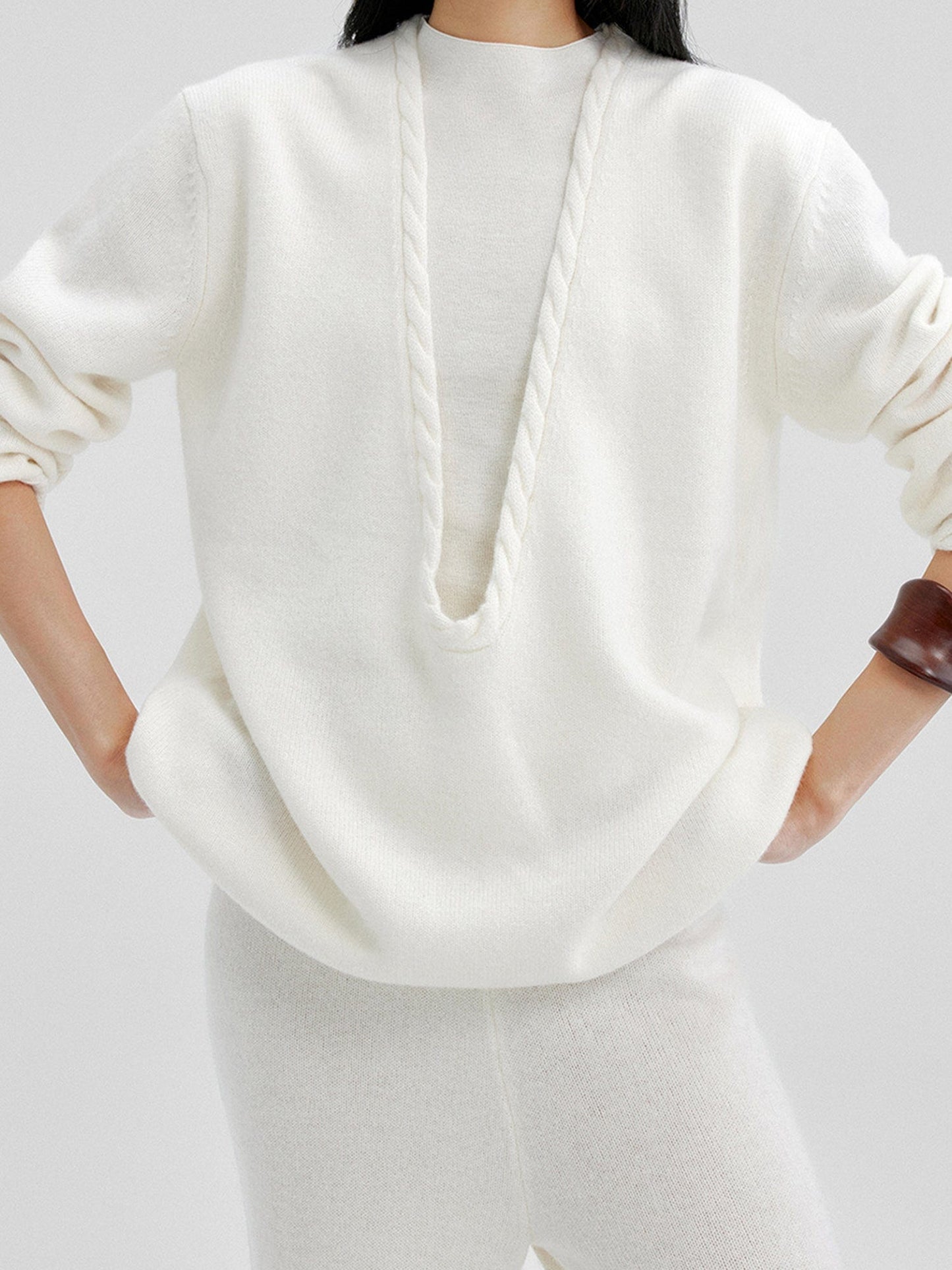 Woman wearing Arkin Studio cable knit v-neck wool sweater in white with a white top inside and matching white knit pants, both hands on hips, standing facing the camera, on a white background