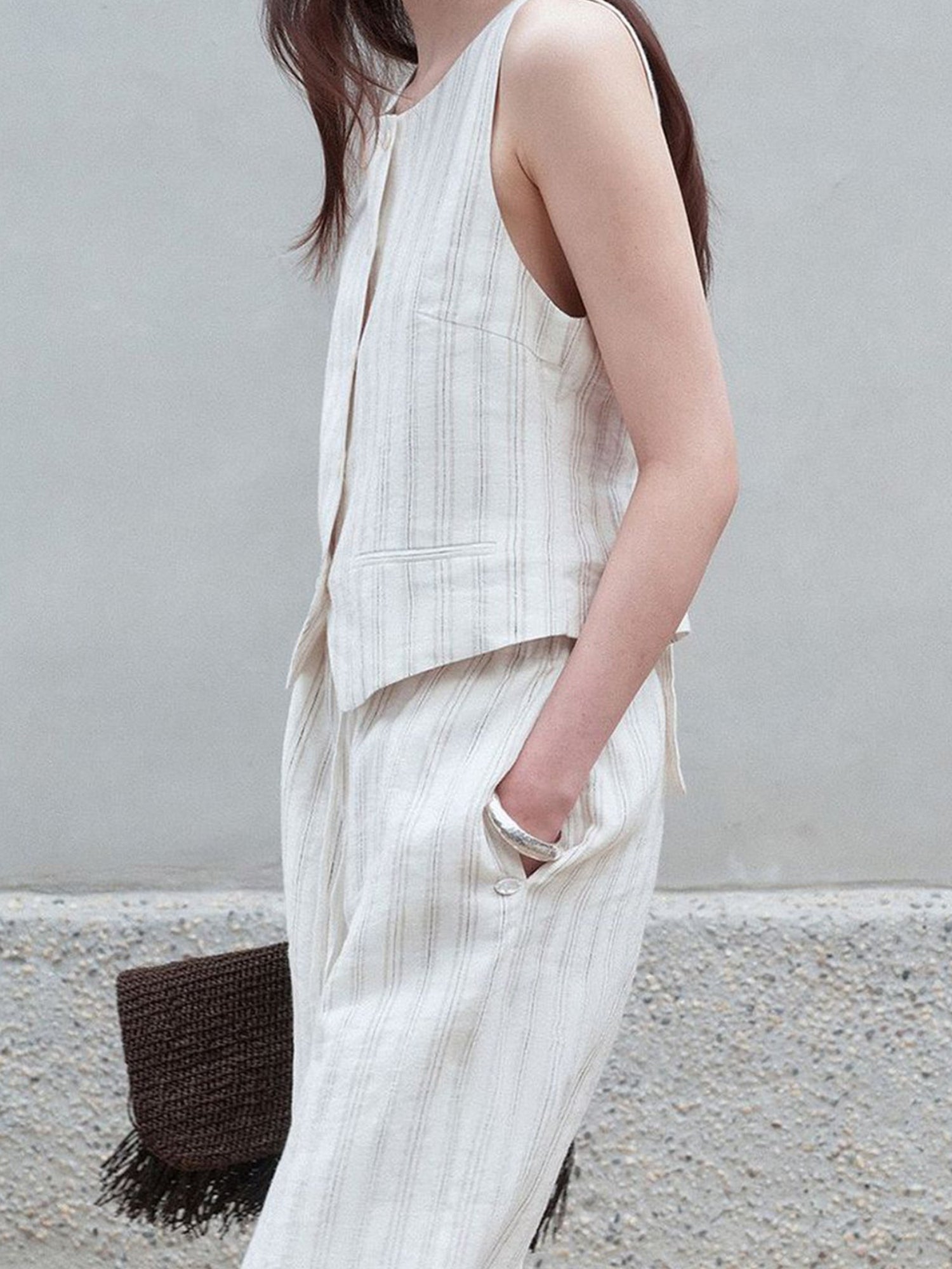 Woman wearing Arkin Studio classic striped vest in ivory and striped trousers in ivory, a brown handbag in her right hand, left hand in the trousers‘ pocket, standing sideways, a wall behind her
