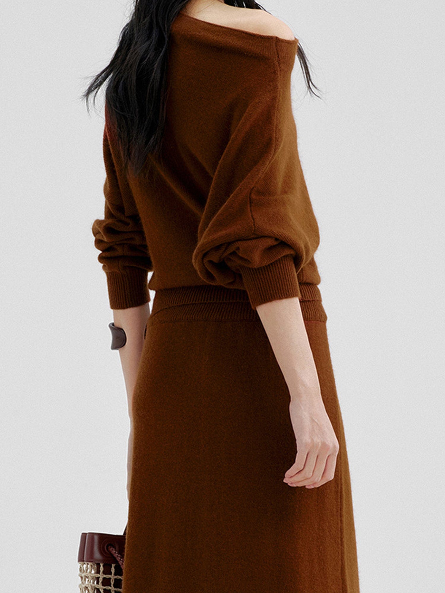 Woman wearing Arkin Studio cold shoulder slim knit top in brown and matching skirt, holding a brown bag, standing facing a white wall, on a white background