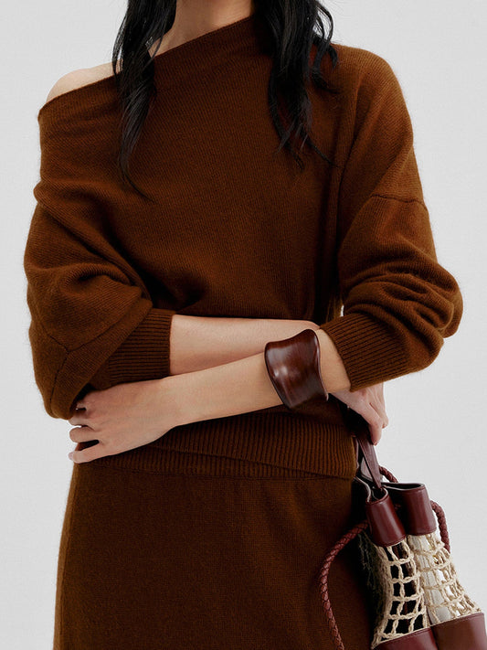 Woman wearing Arkin Studio cold shoulder slim knit top in brown and matching skirt, both arms crossing over the chest, holding a brown bag, standing facing the camera, on a white background