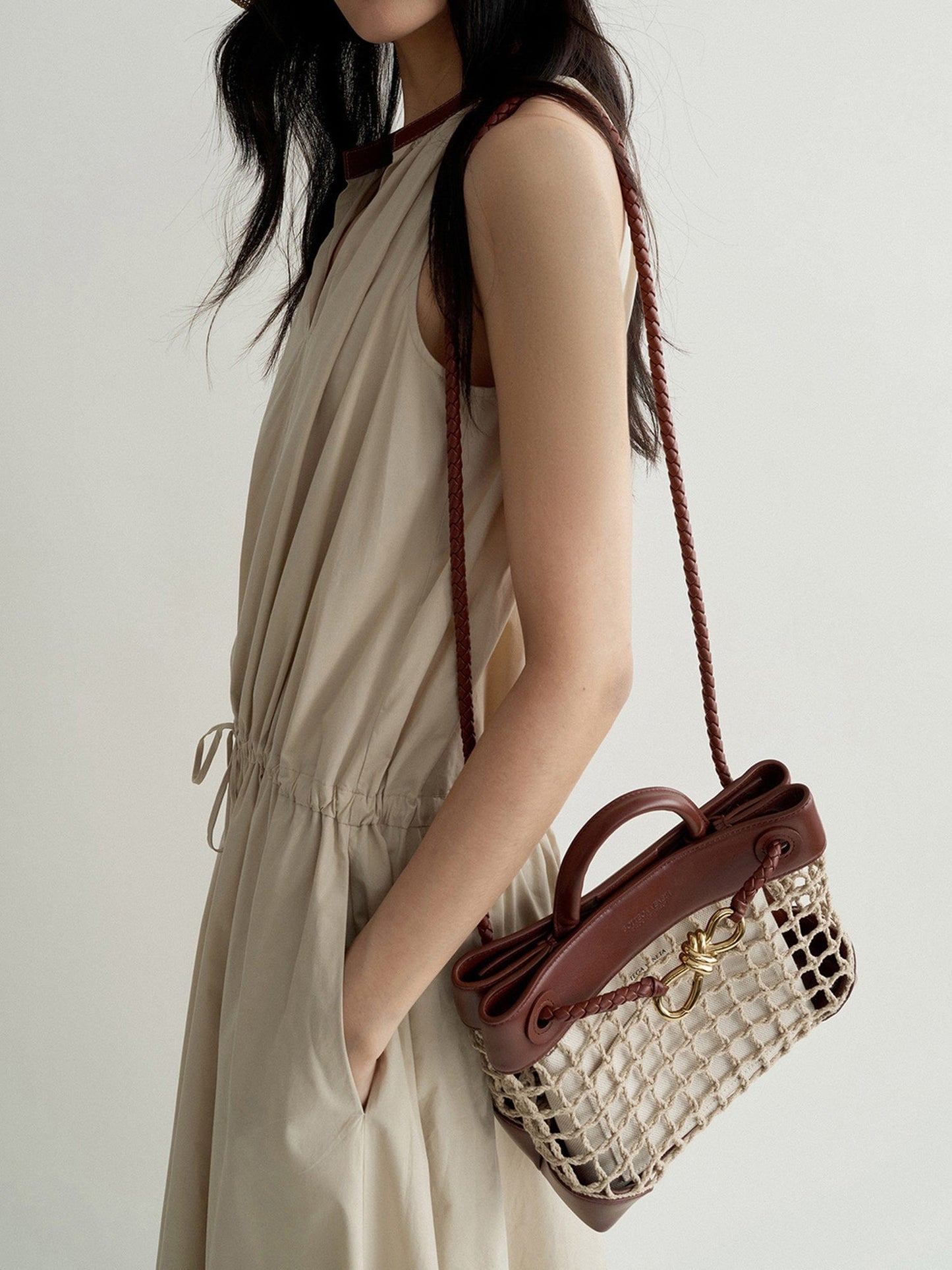 Woman wearing Arkin Studio cotton shift dress with leather collar in cream, with a brown bag on her left shoulder, one hand in the pocket of the dress, standing sideways, on a white background
