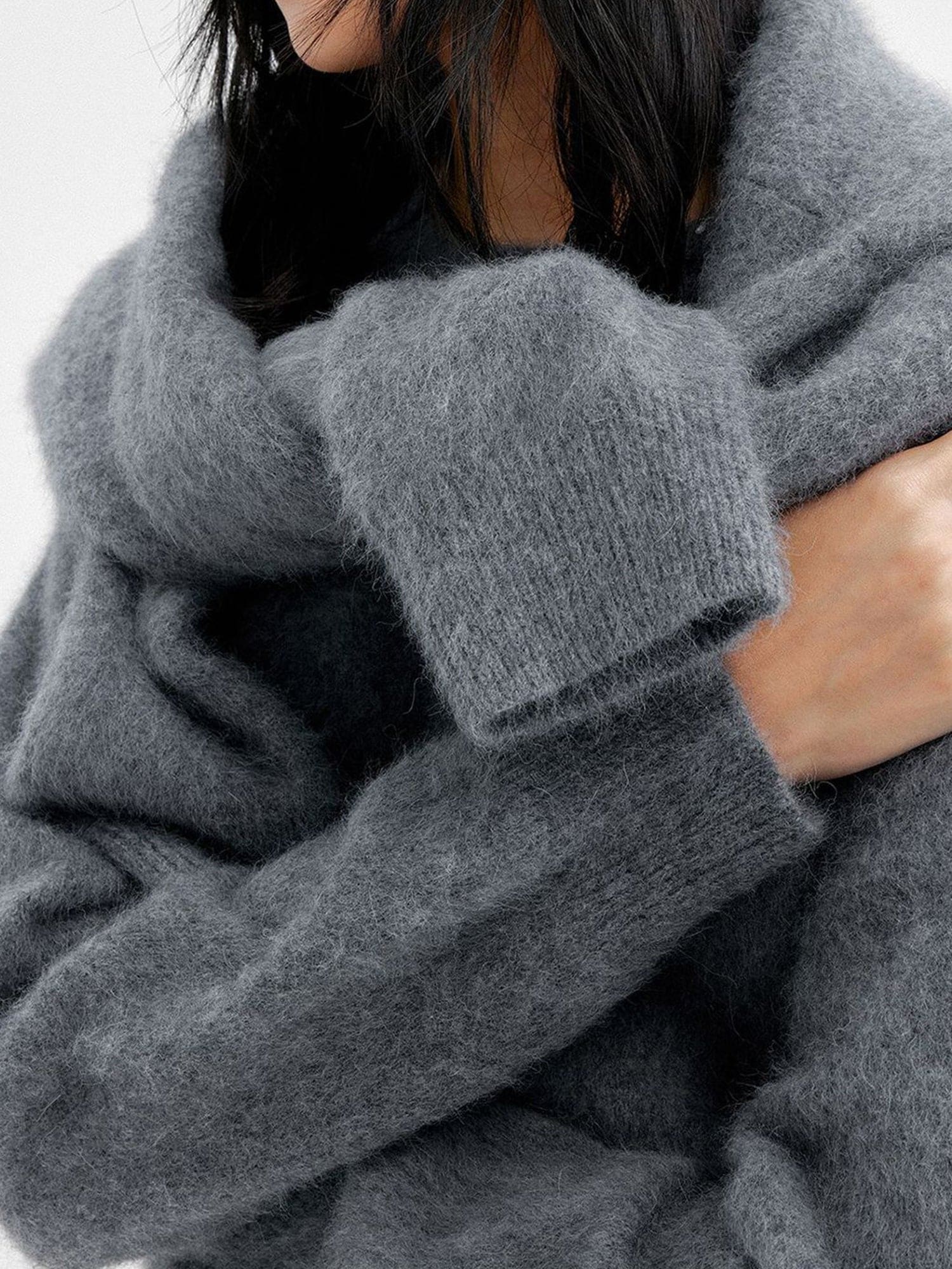 Close up of a woman wearing Arkin Studio cozy crewneck wool sweater in charcoal with a charcoal knitwear on her back, focus on both arms in crossing over her chest, standing sideways, on a white background