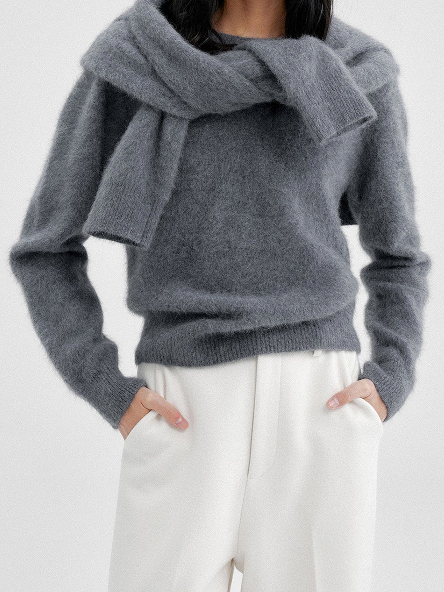 Woman wearing Arkin Studio cozy crewneck wool sweater in charcoal with a charcoal knitwear on her back and matching white pants, both hands in pockets, standing facing the camera, on a white background