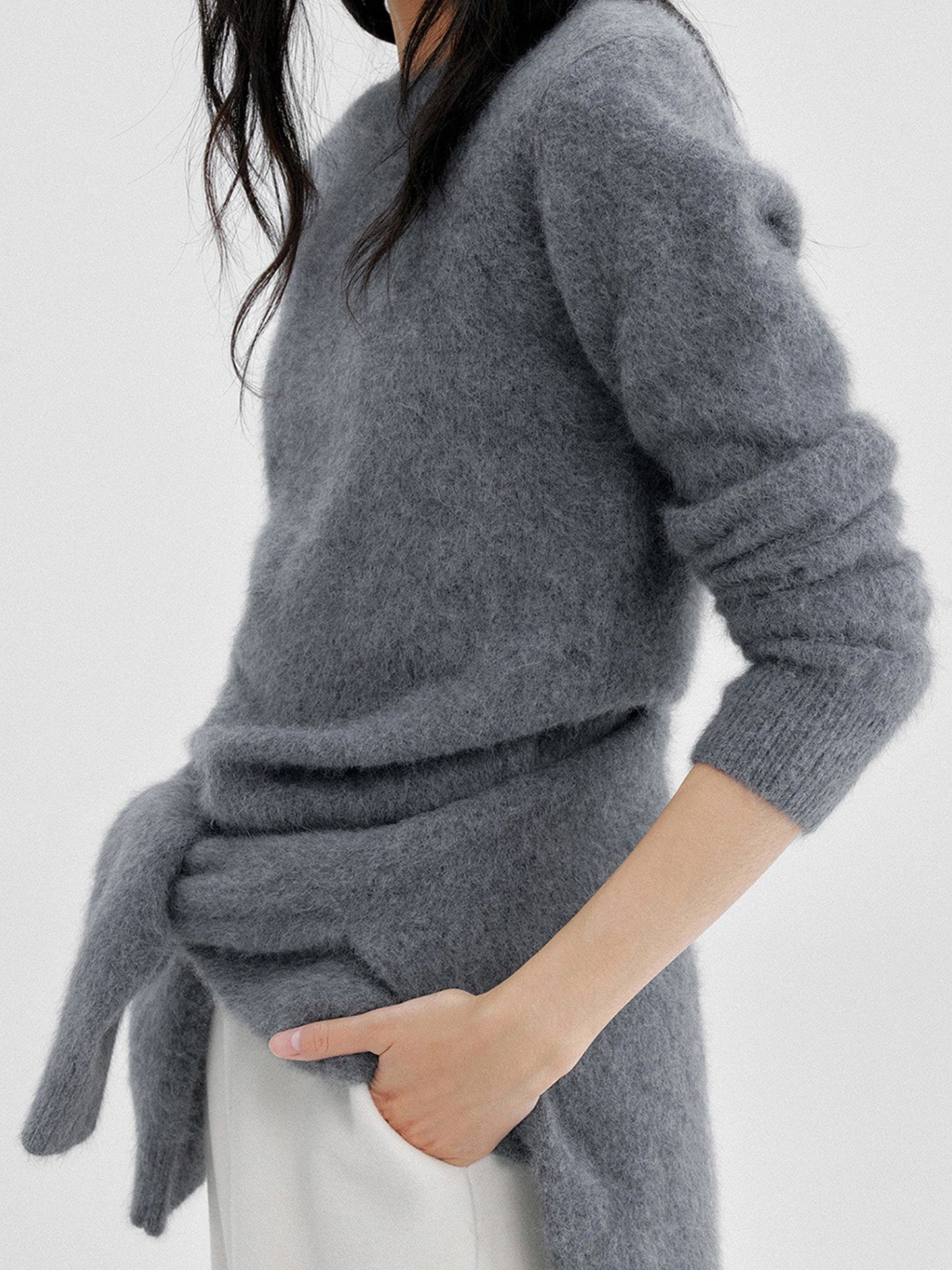 Woman wearing Arkin Studio cozy crewneck wool sweater in charcoal with a charcoal knitwear on her back and matching white pants , both hands in pockets, standing sideways, on a white background