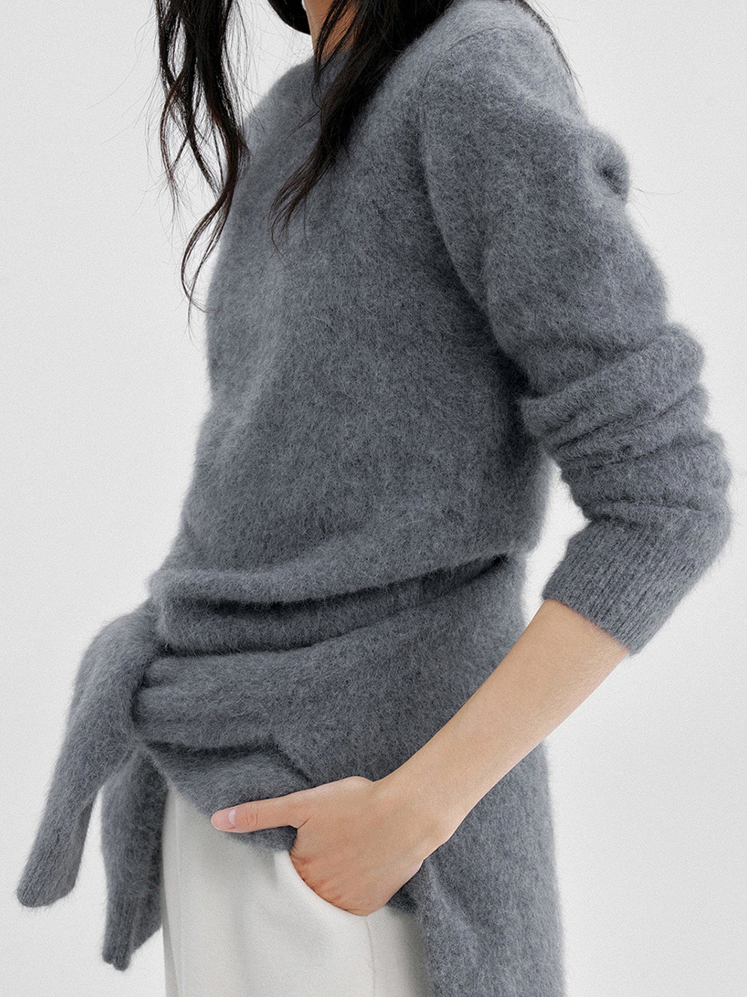 Woman wearing Arkin Studio cozy crewneck wool sweater in charcoal with a charcoal knitwear on her back and matching white pants , both hands in pockets, standing sideways, on a white background