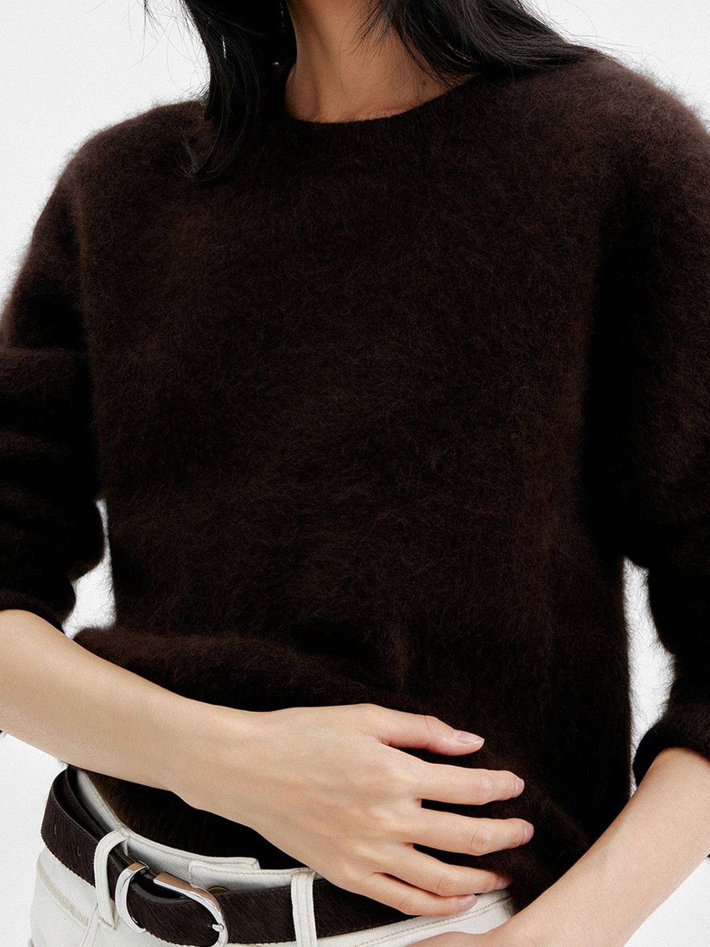 Close up of a woman wearing Arkin Studio cozy crewneck wool sweater in dark-brown and matching white pants with a dark brown belt, focus on the upper part of her body, one hand touching her abdomen, standing facing the camera, on a white background