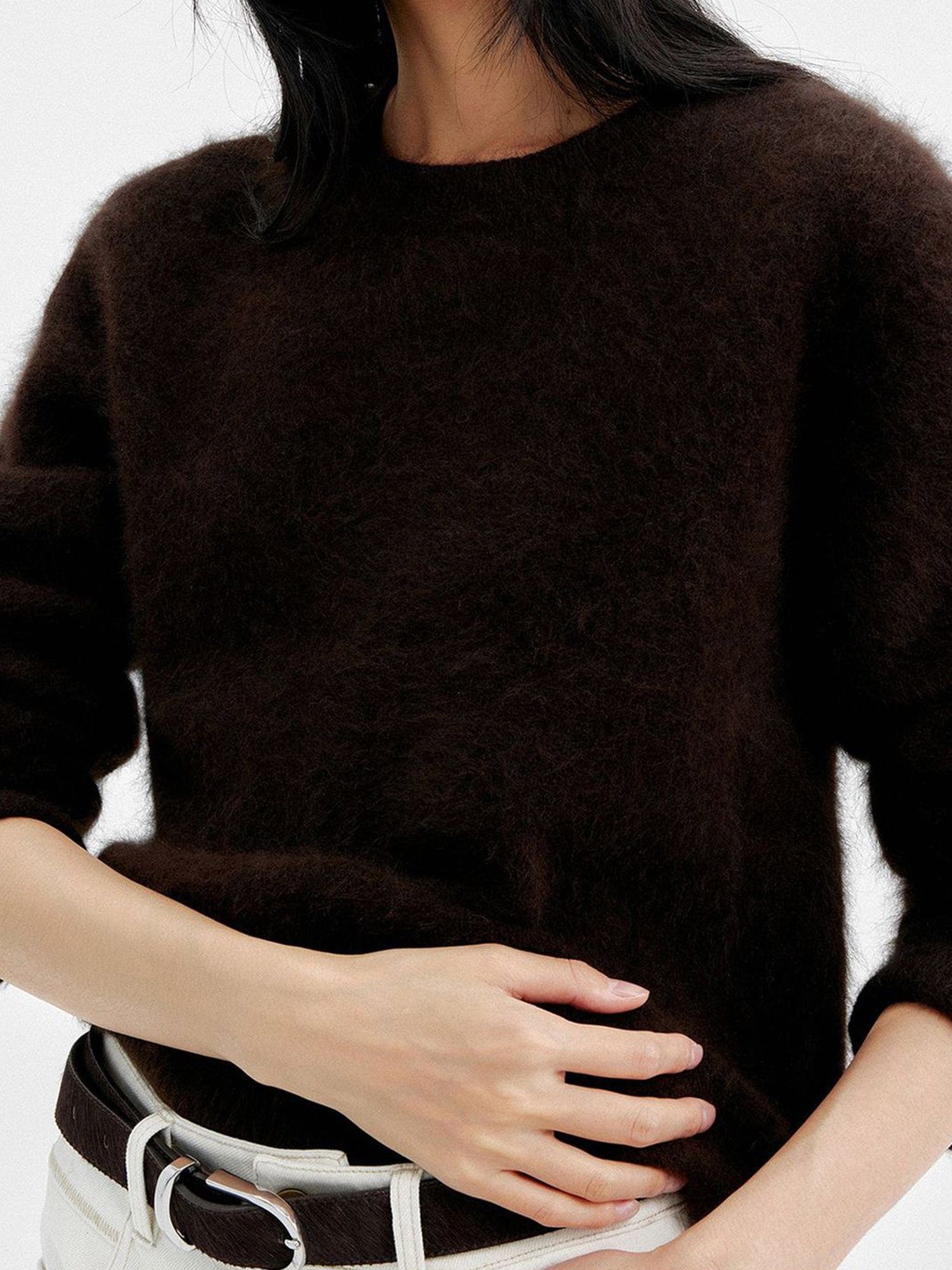 Close up of a woman wearing Arkin Studio cozy crewneck wool sweater in dark-brown and matching white pants with a dark brown belt, focus on the upper part of her body, one hand touching her abdomen, standing facing the camera, on a white background