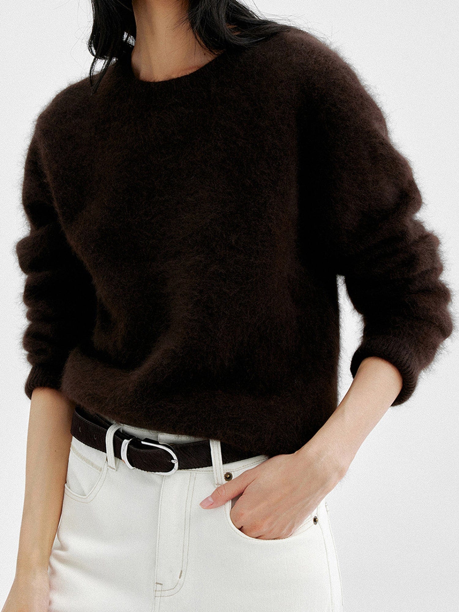 Woman wearing Arkin Studio cozy crewneck wool sweater in dark-brown and matching white pants with a dark brown belt, one hand in a pocket, standing facing the camera, on a white background