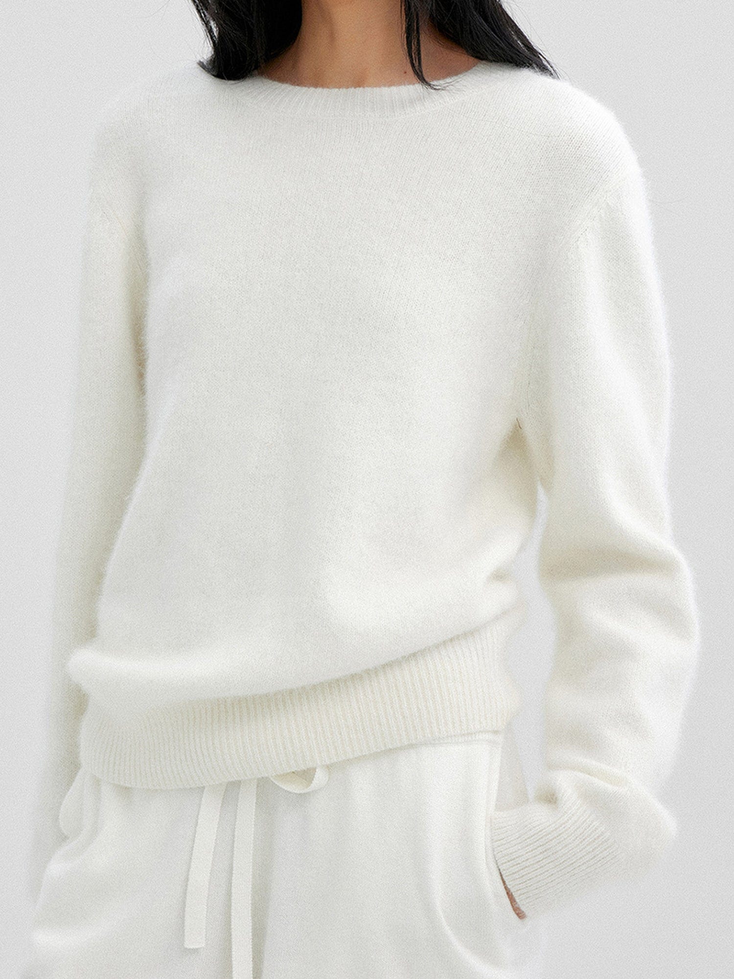 Woman wearing Arkin Studio cozy crewneck wool sweater in ivory and matching shorts, both hands in pockets, standing facing the camera, on a white background