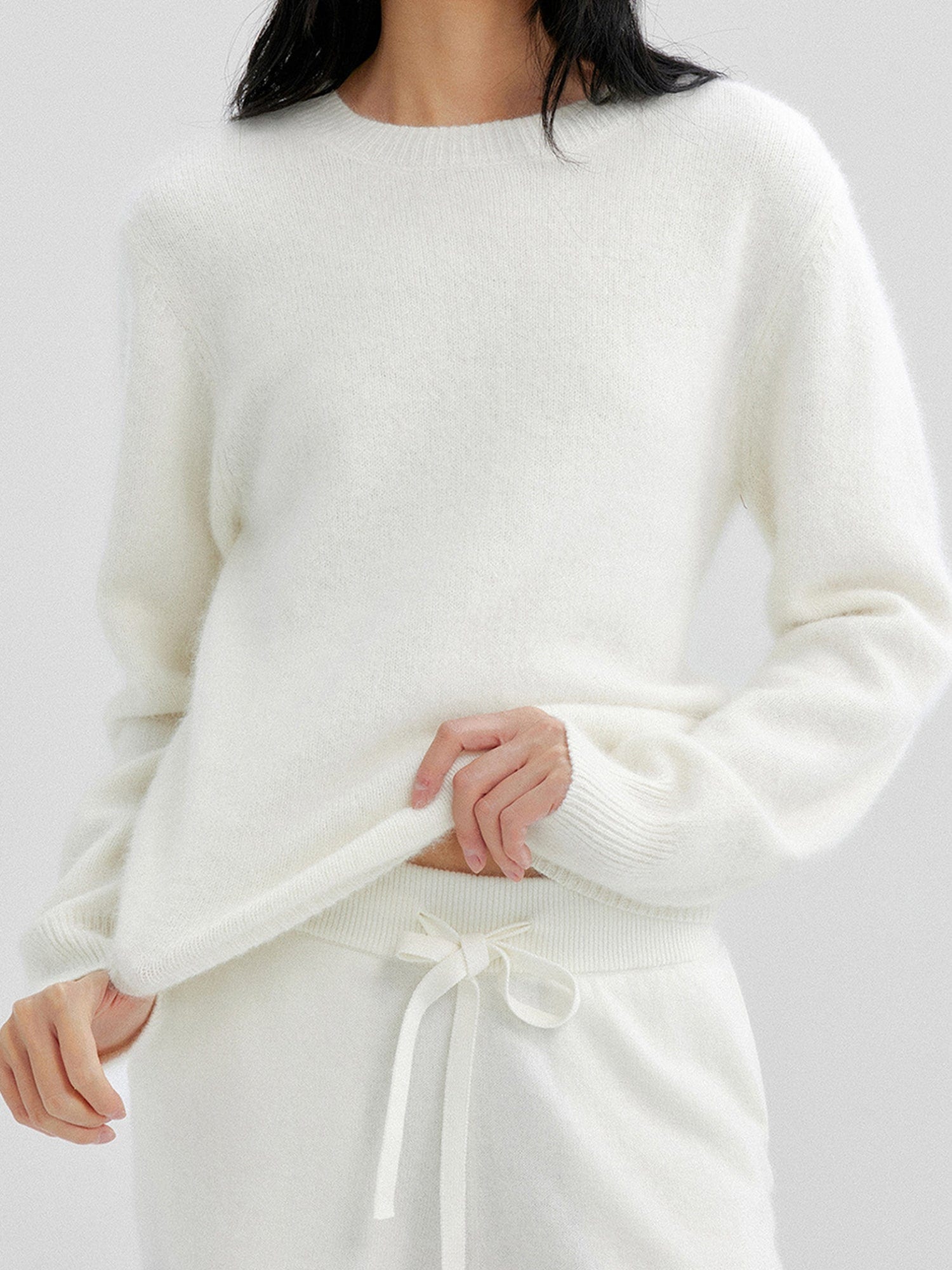 Woman wearing Arkin Studio cozy crewneck wool sweater in ivory and matching shorts, pulling the hem of the sweater with both hands, standing facing the camera, on a white background
