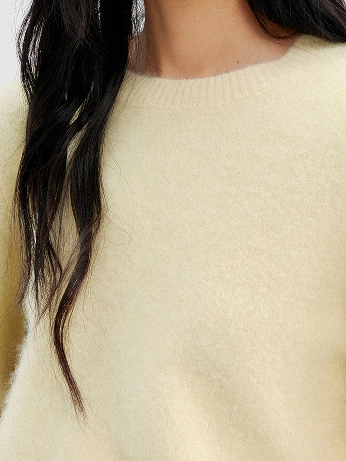 Close up of a woman wearing Arkin Studio cozy crewneck wool sweater in light-yellow, focus on her chest with long hair, standing facing the camera, on a white background