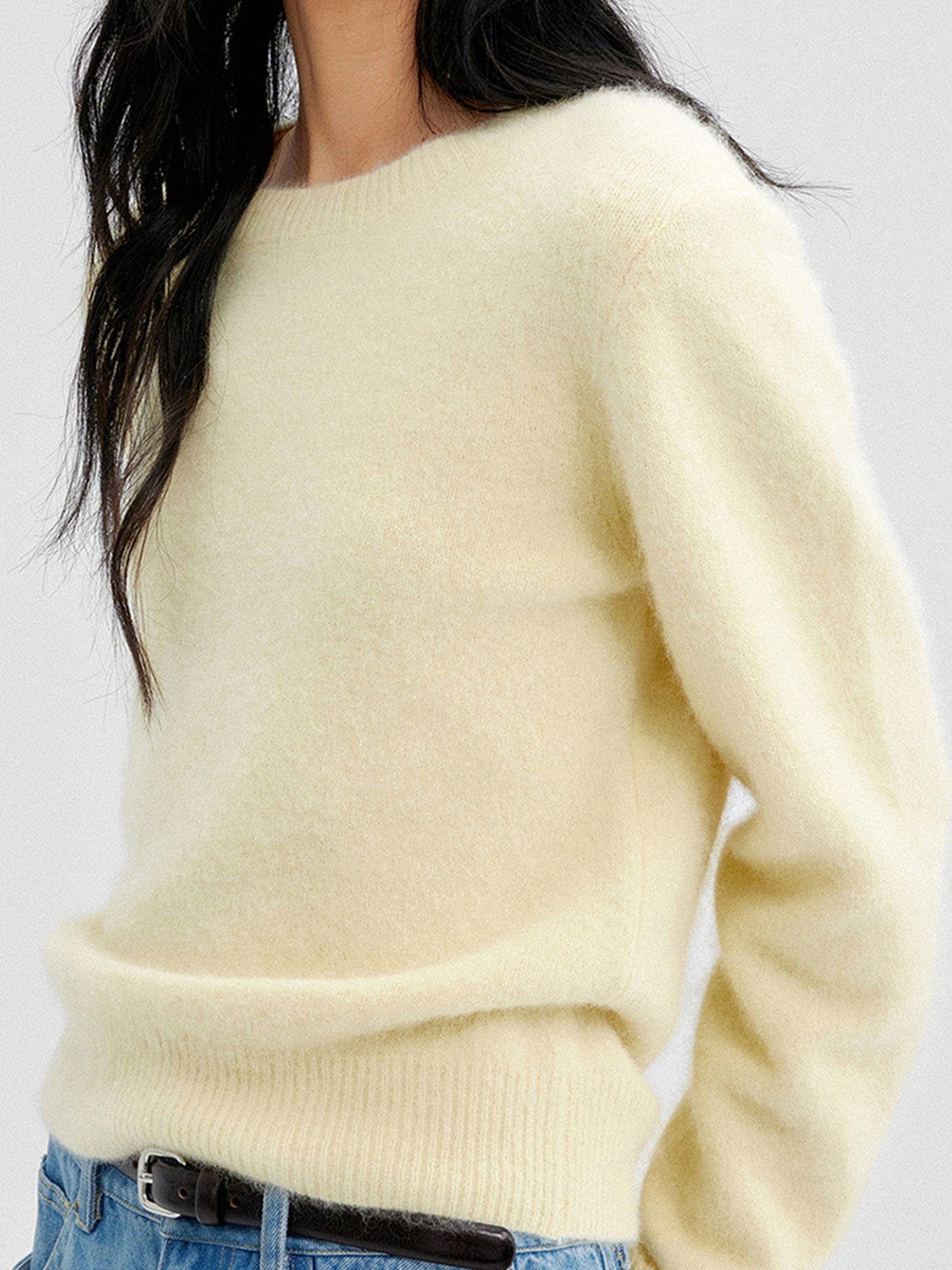 Close up of a woman wearing Arkin Studio cozy crewneck wool sweater in light-yellow and matching blue pants, both hands behind her back, focus on the upper part of her, standing sideways, on a white background