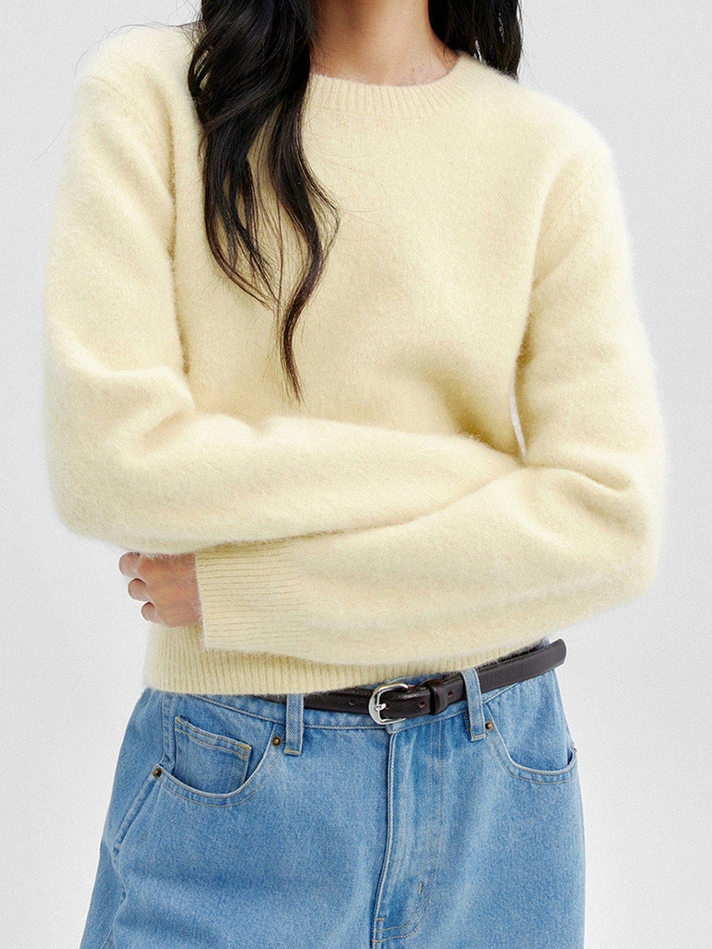 Woman wearing Arkin Studio cozy crewneck wool sweater in light-yellow and  matching blue pants with a black belt, both arms crossing over her abdomen, standing facing the camera, on a white background