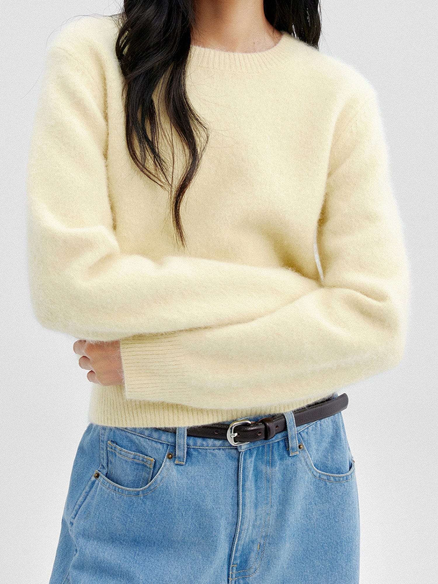 Woman wearing Arkin Studio cozy crewneck wool sweater in light-yellow and  matching blue pants with a black belt, both arms crossing over her abdomen, standing facing the camera, on a white background